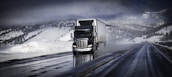 A large truck is driving down a wet highway, surrounded by snow-covered mountains and dark, cloudy skies. The road appears shiny from moisture, likely from melting snow, and the landscape is wintry with snow blanketing the ground and trees.