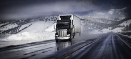 A large truck is driving down a wet highway, surrounded by snow-covered mountains and dark, cloudy skies. The road appears shiny from moisture, likely from melting snow, and the landscape is wintry with snow blanketing the ground and trees.