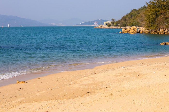 A serene beach scene with golden sand stretching towards the turquoise sea. Gentle waves lap at the shore, and in the distance, sailboats can be seen on the water. The backdrop includes lush green hills and a rocky outcrop.