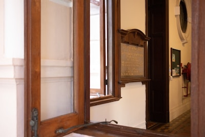 A wooden-framed window is open, offering a view of an interior room with cream-colored walls. To the right of the window, there is a mounted wooden plaque with inscriptions. Below the plaque, a dark door stands ajar next to a cork board with papers pinned to it. A red fire hose is coiled further along the hallway, and geometric patterned tiles cover the floor.