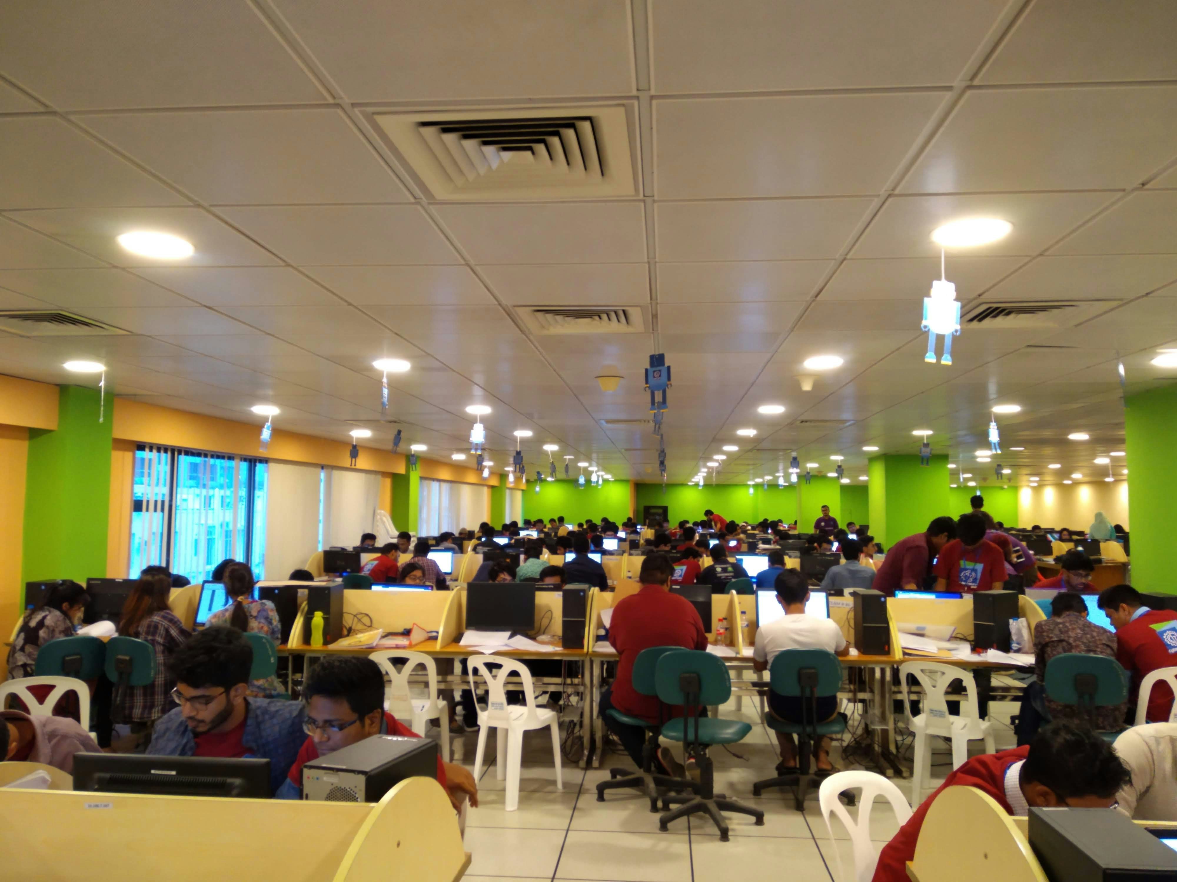 A large room filled with many people seated at long tables, working on computers. The room is brightly lit with overhead lights and decorated with colorful walls, predominantly green and yellow. The setting appears to be a busy office or call center, with a modern suspended ceiling and some hanging decor.