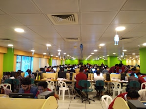 A large room filled with many people seated at long tables, working on computers. The room is brightly lit with overhead lights and decorated with colorful walls, predominantly green and yellow. The setting appears to be a busy office or call center, with a modern suspended ceiling and some hanging decor.
