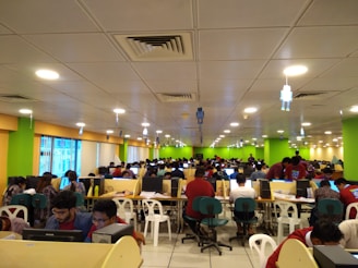 A large room filled with many people seated at long tables, working on computers. The room is brightly lit with overhead lights and decorated with colorful walls, predominantly green and yellow. The setting appears to be a busy office or call center, with a modern suspended ceiling and some hanging decor.