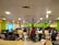 A large room filled with many people seated at long tables, working on computers. The room is brightly lit with overhead lights and decorated with colorful walls, predominantly green and yellow. The setting appears to be a busy office or call center, with a modern suspended ceiling and some hanging decor.