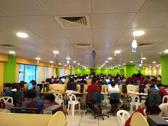 A large room filled with many people seated at long tables, working on computers. The room is brightly lit with overhead lights and decorated with colorful walls, predominantly green and yellow. The setting appears to be a busy office or call center, with a modern suspended ceiling and some hanging decor.