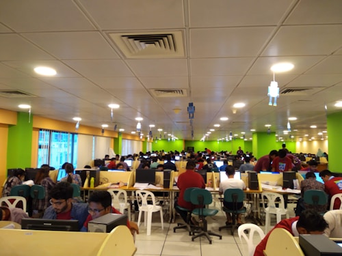 A large room filled with many people seated at long tables, working on computers. The room is brightly lit with overhead lights and decorated with colorful walls, predominantly green and yellow. The setting appears to be a busy office or call center, with a modern suspended ceiling and some hanging decor.