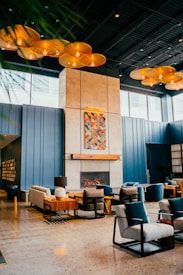 A modern interior with blue paneled walls and a tall fireplace centerpiece featuring a colorful abstract painting above it. The space is furnished with cushioned chairs and sofas arranged around a low wooden table. Overhead, a series of warm, circular light fixtures provide a soft ambiance. The floor is polished concrete, enhancing the sleek and contemporary aesthetic.