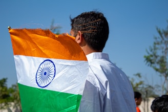 A person with short black hair is holding an Indian flag with an orange, white, and green tricolor and a blue Ashoka Chakra in the center. The individual is wearing a white shirt and standing outdoors on a sunny day with a clear blue sky and trees in the background.