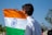 A person with short black hair is holding an Indian flag with an orange, white, and green tricolor and a blue Ashoka Chakra in the center. The individual is wearing a white shirt and standing outdoors on a sunny day with a clear blue sky and trees in the background.