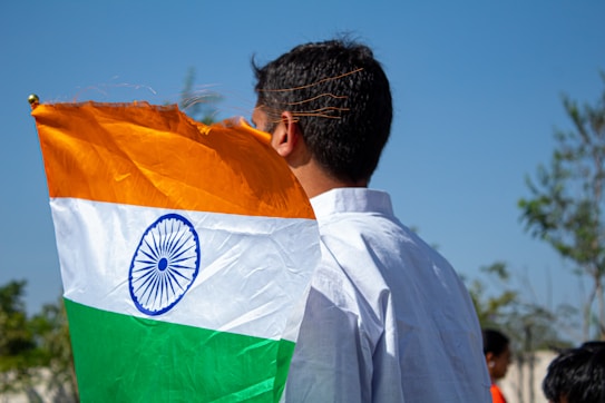 A person with short black hair is holding an Indian flag with an orange, white, and green tricolor and a blue Ashoka Chakra in the center. The individual is wearing a white shirt and standing outdoors on a sunny day with a clear blue sky and trees in the background.