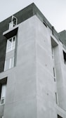 A modern, multi-story building with a minimalist design featuring geometric shapes and parallel lines. The structure showcases a combination of smooth, light gray concrete surfaces and large windows, some of which are open. The building's design is simple yet striking, emphasizing clean lines and functional architecture.