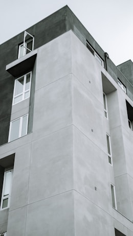 A modern, multi-story building with a minimalist design featuring geometric shapes and parallel lines. The structure showcases a combination of smooth, light gray concrete surfaces and large windows, some of which are open. The building's design is simple yet striking, emphasizing clean lines and functional architecture.