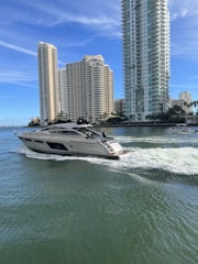 A modern yacht cruises through calm water with a backdrop of tall, luxurious high-rise buildings near the shoreline. The sky is clear with a few wispy clouds, and the water shows gentle ripples caused by the yacht's movement.