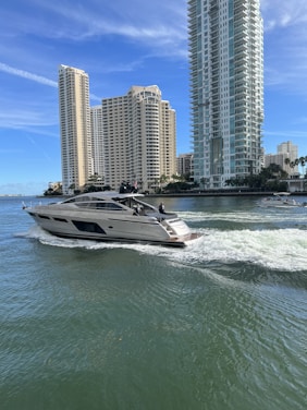 A modern yacht cruises through calm water with a backdrop of tall, luxurious high-rise buildings near the shoreline. The sky is clear with a few wispy clouds, and the water shows gentle ripples caused by the yacht's movement.