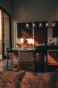A warm, ambient dining area featuring a wooden table surrounded by chairs. The lighting fixture above the table gives a soft glow, and there are decorative panels on the wall. Sunlight filters through a sliding glass door, casting shadows and highlighting the rich wooden textures.
