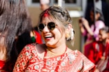 A woman dressed in a traditional embroidered sari is smiling joyfully, wearing sunglasses and gold jewelry. Her hair is styled elegantly with white flowers, and she is surrounded by people in similar red attire.