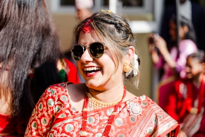 A woman dressed in a traditional embroidered sari is smiling joyfully, wearing sunglasses and gold jewelry. Her hair is styled elegantly with white flowers, and she is surrounded by people in similar red attire.