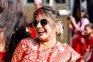A woman dressed in a traditional embroidered sari is smiling joyfully, wearing sunglasses and gold jewelry. Her hair is styled elegantly with white flowers, and she is surrounded by people in similar red attire.