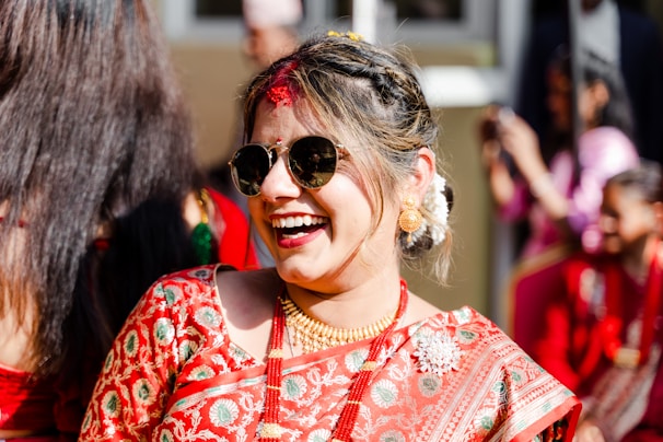 A woman dressed in a traditional embroidered sari is smiling joyfully, wearing sunglasses and gold jewelry. Her hair is styled elegantly with white flowers, and she is surrounded by people in similar red attire.
