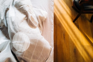 A soft, crumpled blanket lies on a bed with a checkered pattern. Sunlight streams in, casting warm, golden light and shadows across a wooden floor.