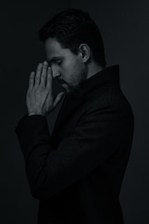 A person with dark hair appears in a pensive pose, with hands clasped in front of their face, eyes closed, and a thoughtful expression. The lighting is low, creating a moody and introspective atmosphere.
