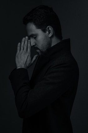 A person with dark hair appears in a pensive pose, with hands clasped in front of their face, eyes closed, and a thoughtful expression. The lighting is low, creating a moody and introspective atmosphere.