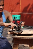 A person wearing a blue tank top is using a green and black electric router to carve a piece of circular wood. The wood is placed on a small wooden stool. The background consists of an orange wall and a yellow cord is visible.