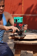 A person wearing a blue tank top is using a green and black electric router to carve a piece of circular wood. The wood is placed on a small wooden stool. The background consists of an orange wall and a yellow cord is visible.