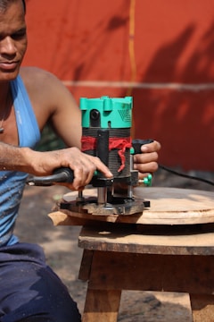 A person wearing a blue tank top is using a green and black electric router to carve a piece of circular wood. The wood is placed on a small wooden stool. The background consists of an orange wall and a yellow cord is visible.
