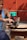 A person wearing a blue tank top is using a green and black electric router to carve a piece of circular wood. The wood is placed on a small wooden stool. The background consists of an orange wall and a yellow cord is visible.