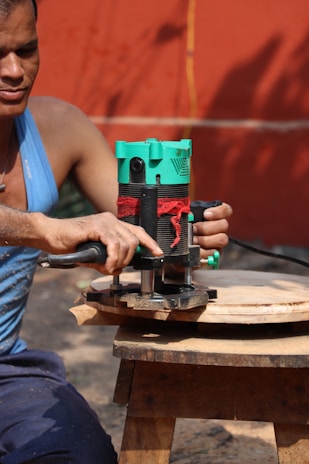 A person wearing a blue tank top is using a green and black electric router to carve a piece of circular wood. The wood is placed on a small wooden stool. The background consists of an orange wall and a yellow cord is visible.