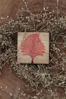 A wooden surface features a small square plaque with a vibrant red tree design. Thin, dry branches with small leaves encircle the plaque, adding a rustic and natural frame to the artwork.