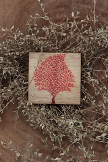 A wooden surface features a small square plaque with a vibrant red tree design. Thin, dry branches with small leaves encircle the plaque, adding a rustic and natural frame to the artwork.