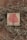 A wooden surface features a small square plaque with a vibrant red tree design. Thin, dry branches with small leaves encircle the plaque, adding a rustic and natural frame to the artwork.