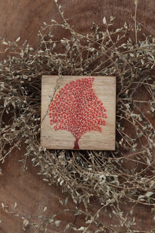 A wooden surface features a small square plaque with a vibrant red tree design. Thin, dry branches with small leaves encircle the plaque, adding a rustic and natural frame to the artwork.