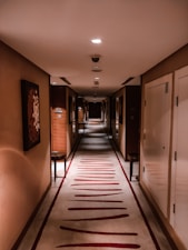 A long and narrow hotel corridor with a patterned carpet and warm lighting. The walls are adorned with abstract artwork, and there are several closed doors on each side with door numbers. The atmosphere is quiet, and the end of the hallway is dimly lit.