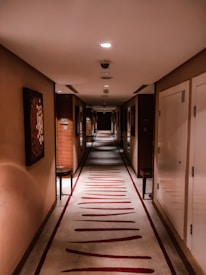 A long and narrow hotel corridor with a patterned carpet and warm lighting. The walls are adorned with abstract artwork, and there are several closed doors on each side with door numbers. The atmosphere is quiet, and the end of the hallway is dimly lit.