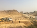 A construction site in a dusty environment with a backhoe loader and a truck working. Several people are standing around the machinery, and a rough dirt road leads towards a group of unfinished buildings in the background. There is a large rock formation or hill visible behind the construction activity.