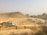 A construction site in a dusty environment with a backhoe loader and a truck working. Several people are standing around the machinery, and a rough dirt road leads towards a group of unfinished buildings in the background. There is a large rock formation or hill visible behind the construction activity.