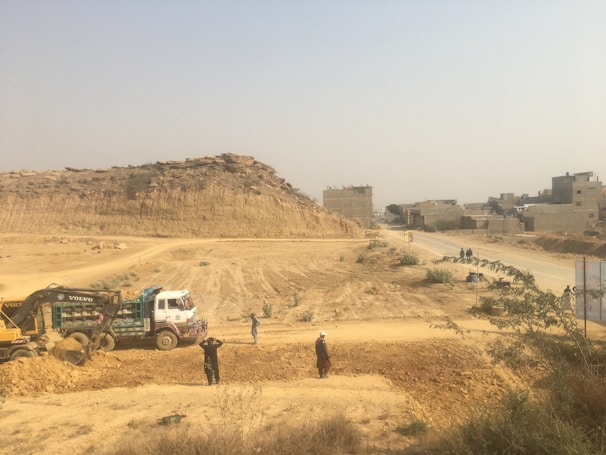 A construction site in a dusty environment with a backhoe loader and a truck working. Several people are standing around the machinery, and a rough dirt road leads towards a group of unfinished buildings in the background. There is a large rock formation or hill visible behind the construction activity.