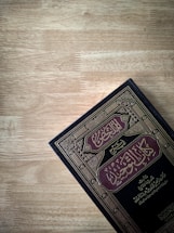 A book with Arabic script on the cover is placed against a wooden surface. The cover features intricate borders and patterns in gold and maroon colors.