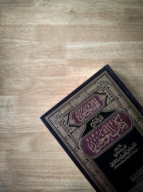 A book with Arabic script on the cover is placed against a wooden surface. The cover features intricate borders and patterns in gold and maroon colors.