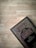 A book with Arabic script on the cover is placed against a wooden surface. The cover features intricate borders and patterns in gold and maroon colors.
