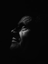 A striking black and white portrait featuring the profile of an elderly person with dramatic lighting enhancing the textures and details of their skin. The contrast creates a profound and introspective atmosphere, highlighting the serene expression.