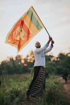 A man stands in a grassy field holding a large flag. The flag has a lion emblem with red, yellow, green, and orange colors. He is wearing a white shirt and long pants with traditional patterns. The background shows a sunset with blurred trees.