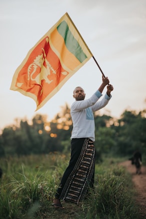 A man stands in a grassy field holding a large flag. The flag has a lion emblem with red, yellow, green, and orange colors. He is wearing a white shirt and long pants with traditional patterns. The background shows a sunset with blurred trees.