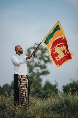 A person stands in a grassy field holding a vibrant flag. The flag is primarily red and yellow, featuring a lion emblem. The individual is dressed in traditional attire, including a white shirt and patterned garment. The sky is a soft blue with some clouds, and there are trees in the background.