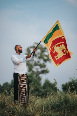 A person stands in a grassy field holding a vibrant flag. The flag is primarily red and yellow, featuring a lion emblem. The individual is dressed in traditional attire, including a white shirt and patterned garment. The sky is a soft blue with some clouds, and there are trees in the background.