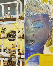 A colorful and eclectic interior with a large mural of a serene Buddha face painted in blue shades. Surrounding the mural, there is a shelf filled with various decorative items including books, plants, tea cups, and figurines. The shelf also features Christmas decorations such as a hanging Santa Claus ornament and tinsels.
