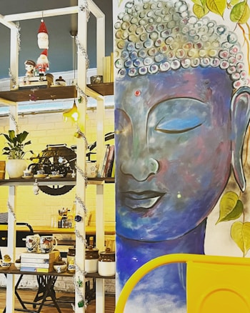 A colorful and eclectic interior with a large mural of a serene Buddha face painted in blue shades. Surrounding the mural, there is a shelf filled with various decorative items including books, plants, tea cups, and figurines. The shelf also features Christmas decorations such as a hanging Santa Claus ornament and tinsels.